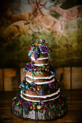 three tier naked wedding cake decorated with purple grape bunches, greenery, strawberries and purple cabbage leaves on top of wooden cake stand