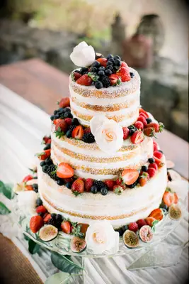 three tier naked wedding cake decorated with powdered sugar, white roses, sliced figs, strawberries and blueberries