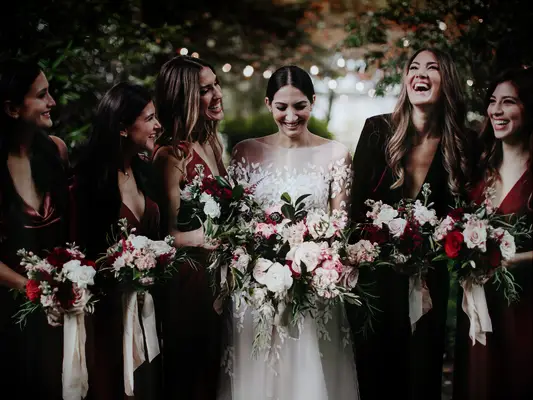 bride stands with bridesmaids wearing red velvet dresses and holding pink and dark burgundy bouquets with long ribbons