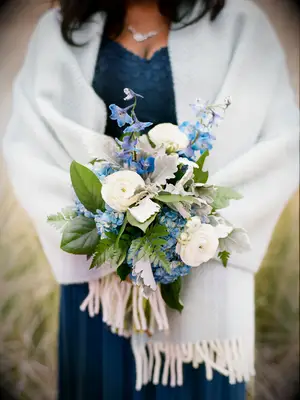 bridesmaid wearing light blue shawl holds small winter bouquet with blue delphinium, white ranunculus and greenery