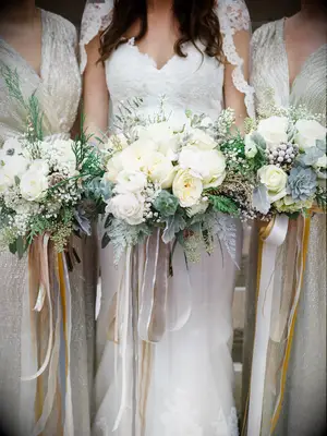 bride and bridesmaids hold bouquets with ivory roses, dusty miller, silver brunia, eucalyptus and long gold and silver metallic ribbons