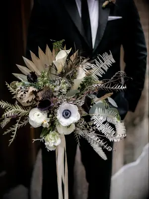 groom holding modern boho winter wedding bouquet with dried fan palm, anemones, ranunculus and greenery in ivory, beige and green tones