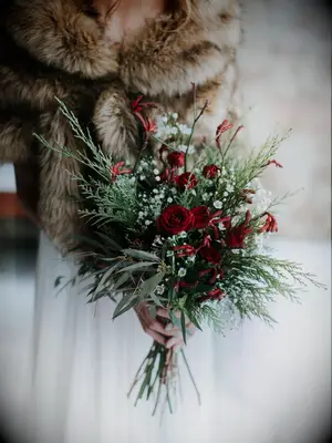 bride wearing brown fur wrap holds winter wedding bouquet with red spray roses and evergreen branches