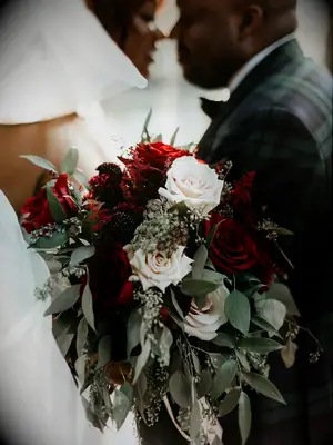 closeup of winter wedding bouquet featuring light pink roses, red roses and greenery with couple kissing in background