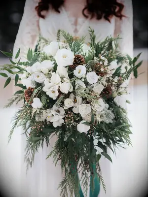 winter wedding bouquet with white flowers, evergreen branches, pinecones and long green ribbons tied to stem