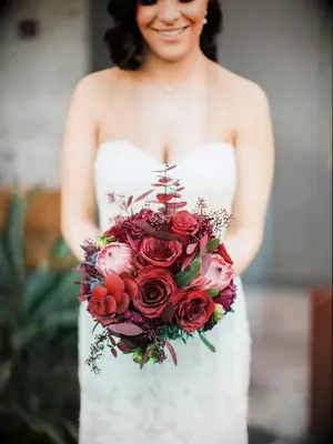 bride holds small rounded wedding bouquet with red roses, proteas and red spray painted greenery