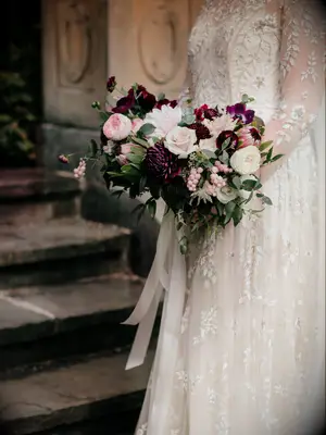 bride wearing long sleeve wedding dress holds winter bouquet of burgundy and light pink flowers with greenery and long ribbons