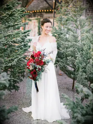 bride standing in an aisle at christmas tree farm holding a red and burgundy cascading wedding bouquet
