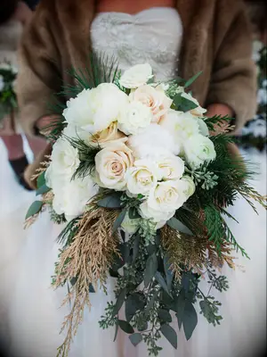 bride wearing brown fur jacket holds winter wedding bouquet with white peonies, blush roses, cotton puffs and gold spray painted greenery