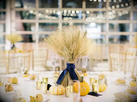 fall wedding centerpiece with wheat stalks tied with navy blue satin bow and orange and white pumpkins around the center of the table