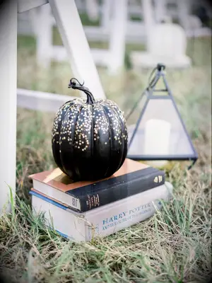 wedding ceremony aisle decor black painted pumpkin with mini gold star glitter sitting on top of a book stack on the ground