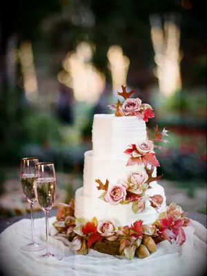 elegant fall wedding cake with white buttercream icing, red fall leaves and mauve roses next to two champagne glasses