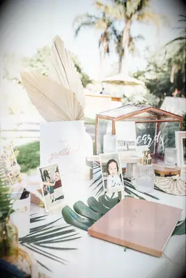 wedding welcome table decorated with gold glass card box, childhood photos of couple, tropical greenery and dried palm leaves