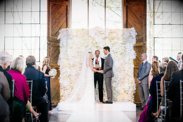 Bride and groom stand in front of a white and pale yellow flower wedding wall during a ceremony.