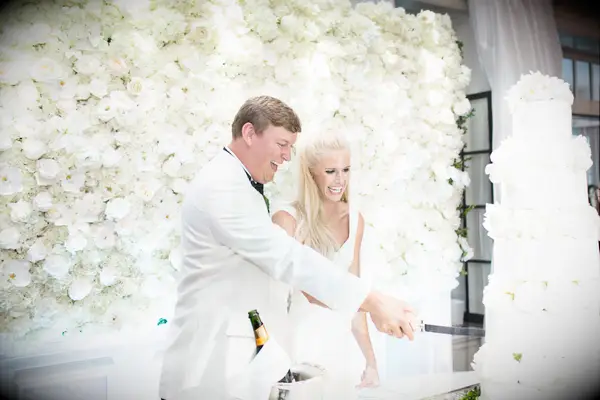 Bride and groom cut their wedding cake with white rose wedding flower wall behind them.