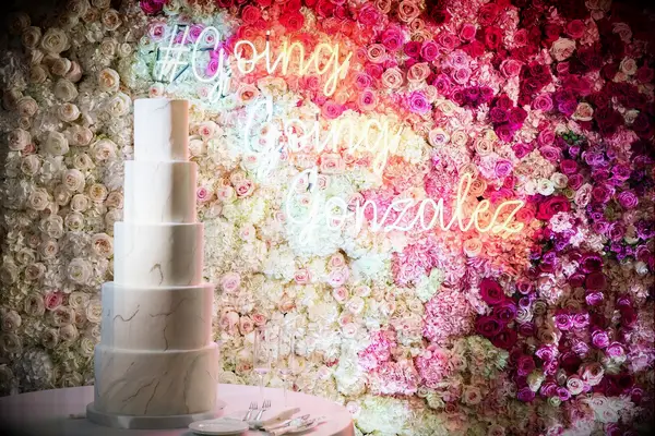 A marble white wedding cake in front of a neon wedding hashtag sign and flower wall. 