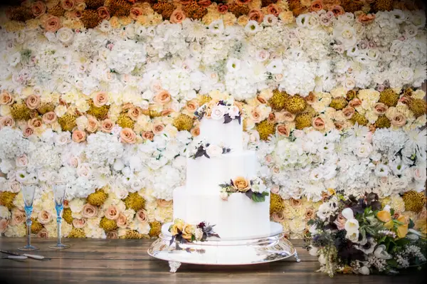 A white wedding cake in front of a yellow and white flower wall.