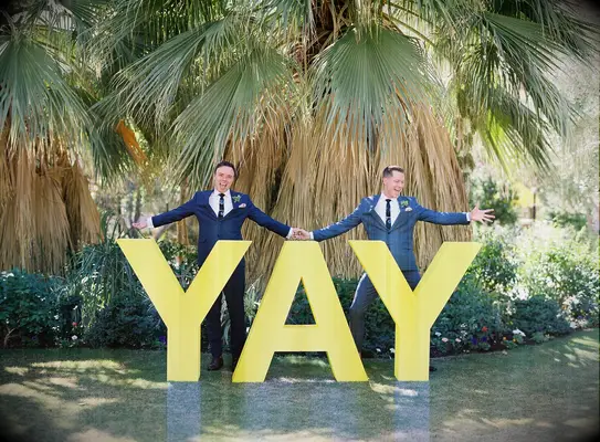 Couple poses with a large sign that reads YAY. 