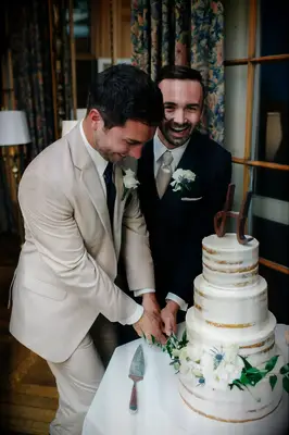 A same-sex couple smiles for the camera while they cut their wedding cake