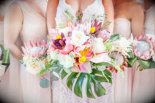 bride and bridesmaid holding tropical wedding bouquets with king proteas, coral charm peonies, craspedia, white roses and monstera leaves