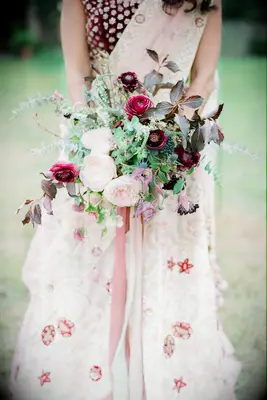 bride in pink and burgundy sari holding fall wedding bouquet with burgundy ranunculus, light pink roses and greenery