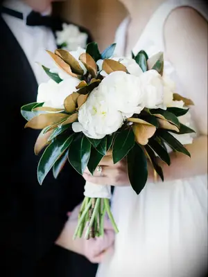 A couple hold a white peony bouquet with green and gold foliage.