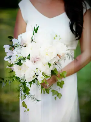 A bride stands holding a burgeoning white peony bouquet.