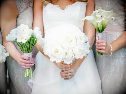 A bride holds a bouquet of white peonies, while her bridesmaids flank her holding complementary white flowers.
