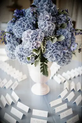 wedding escort card display with pastel blue hydrangeas in a tall white vase surrounded by white tented escort cards
