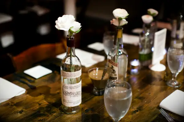 Vintage whiskey bottles hold white roses and sit on a dining table. 