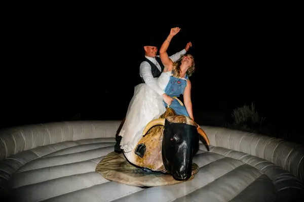Couple rides a mechanical bull while laughing and waving their arms in the air. 