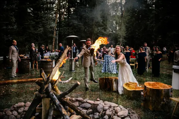 Newlyweds and the wedding party gather around a rustic bonfire in the woods. 