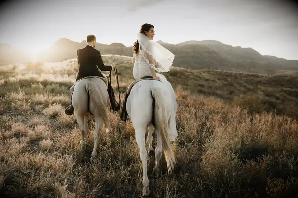 Newlyweds ride horses through a beautiful natural scene. 