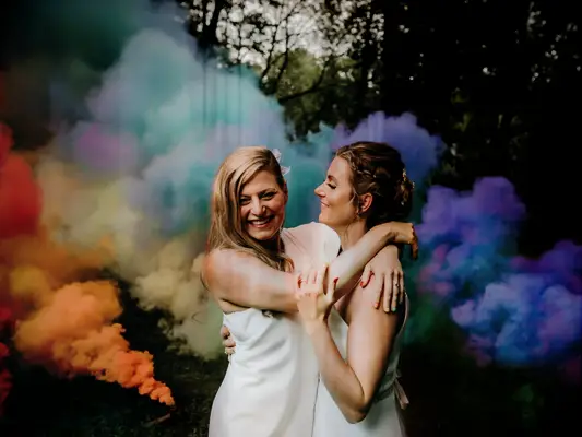 Two brides pose amongst rainbow smoke