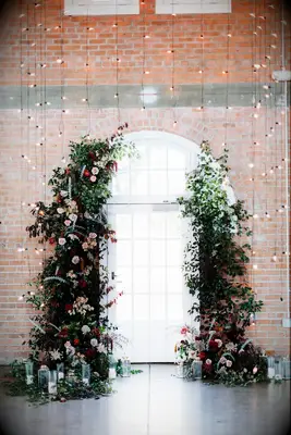 wedding ceremony flower arch in front of string light curtain and brick wall
