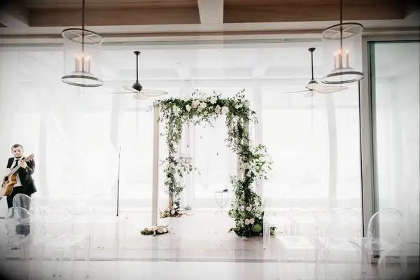Minimal white chuppah with asymmetrical floral decorations surrounded by ghost chairs.