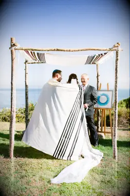 Couple wrapped in prayer shawl underneath birch chuppah.