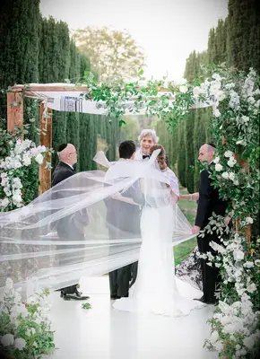 Couple under greenery-covered Chuppah with veil draped over them.