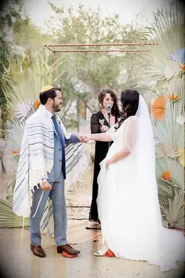 Couple standing in front of copper chuppah decorated with dried palm fronds. 