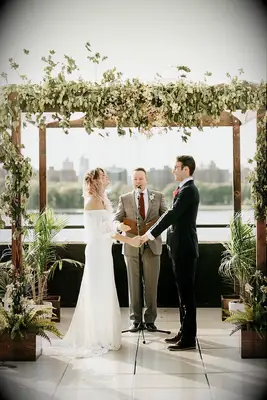 Couple holding hands under wood chuppah decorated with greenery.