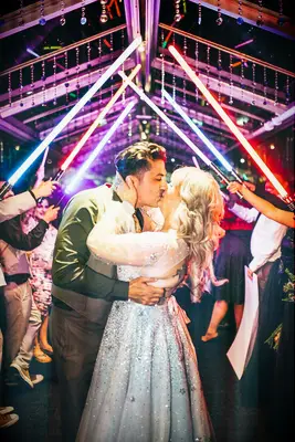 Bride and groom kiss under lightsaber arch.
