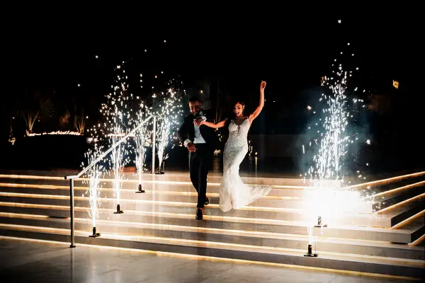 Bride and groom walking down stairs with sparklers machines next to them.
