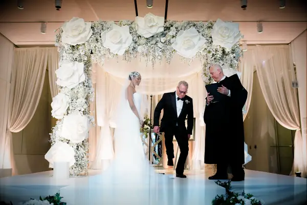 Groom breaking glass during Jewish wedding ceremony under rose-covered chuppah