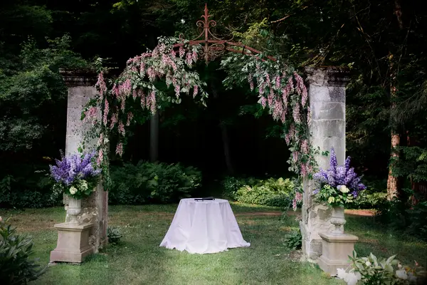 outdoor wedding ceremony altar with stone arch covered in pink wisteria vines with purple larkspur arrangements on both sides