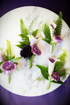 round marble tray showcasing white and purple calla lily boutonnieres with fern leaves and pink roses
