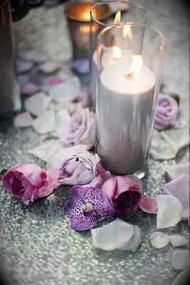 wedding centerpiece with light purple pillar candles in glass vases on silver sequined tablecloth surrounded by purple rose petals and flowers