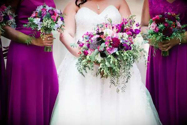 bride holding purple and pink wedding bouquet with protea, dahlias, roses, calla lilies and greenery while standing next to bridesmaids with similar bouquets