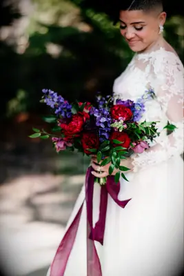 bride holding jewel tone wedding bouquet with red peonies and roses, purple stock flowers, greenery and long fuchsia ribbons tied to the stems