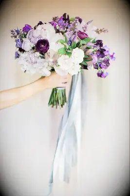 arm extending from the left side of the photo holding purple wedding bouquet against a white backdrop with ranunculus, white peonies, roses, greenery and long blue ribbons tied around the stems