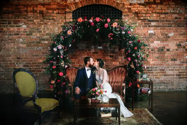 Couple sharing kiss while sitting on velvet couch in front of floral circle arch